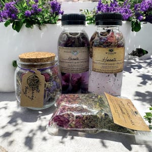 May include: A set of three glass bottles and a jar filled with dried herbs and flowers. The bottles are labeled "Hecate Hand-Curated Liquid Offering Mix" and the jar is labeled "Hand-Crafted Offering Mix".
