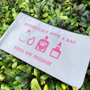May include: A light gray canvas bag with a black zipper, featuring pink text and illustrations. The text reads "DEFINITELY NOT A BAG FULL OF POISON" with images of potion bottles. The bag is set against a backdrop of green foliage.