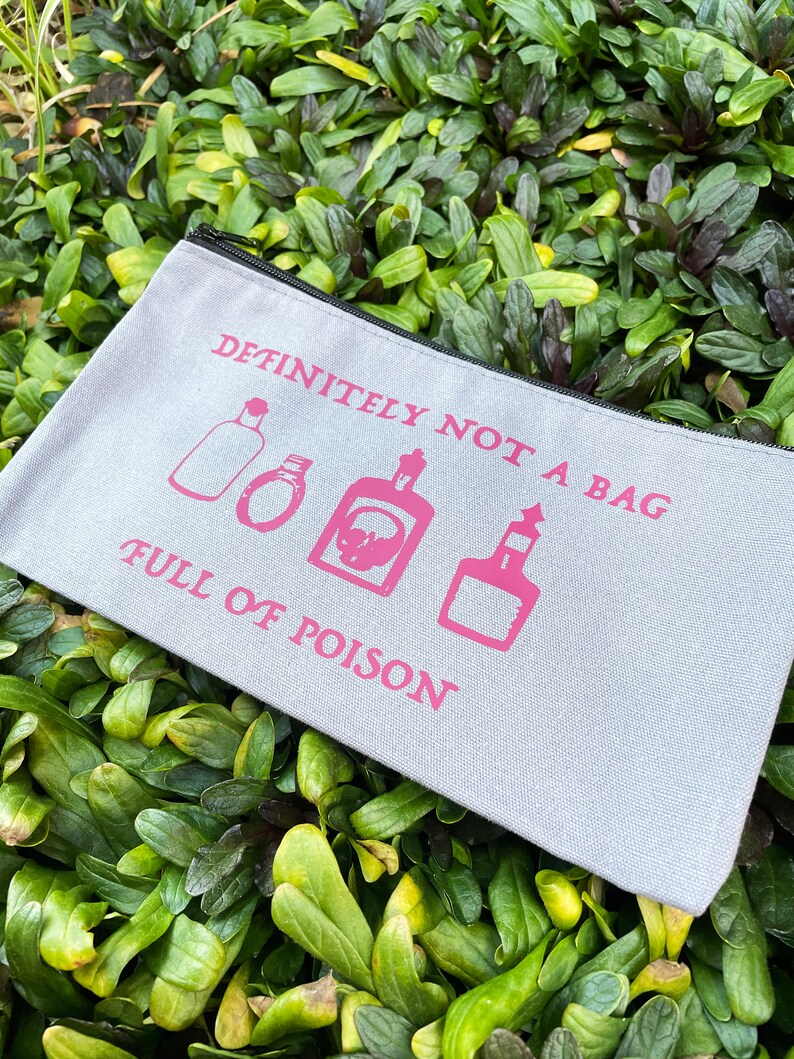 May include: A light gray canvas bag with a black zipper, featuring pink text and illustrations. The text reads "DEFINITELY NOT A BAG FULL OF POISON" with images of potion bottles. The bag is set against a backdrop of green foliage.