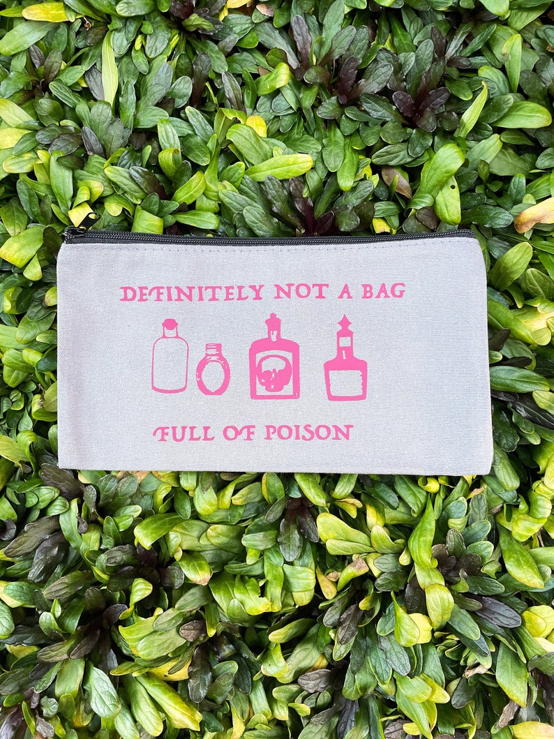 May include: A light gray zippered pouch with the text "DEFINITELY NOT A BAG" and "FULL OF POISON" in pink. The pouch features pink illustrations of potion bottles. The background is a bed of green leaves.