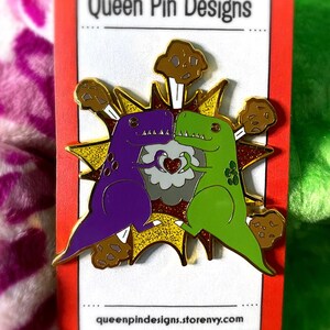 Peut inclure: &Eacute;pingle repr&eacute;sentant deux dinosaures de dessin anim&eacute;, un violet et un vert, s'embrassant dans un motif d'explosion dor&eacute;. L'&eacute;pingle comprend le texte "Queen Pin Designs" et l'adresse queenpindesigns.storenvy.com.