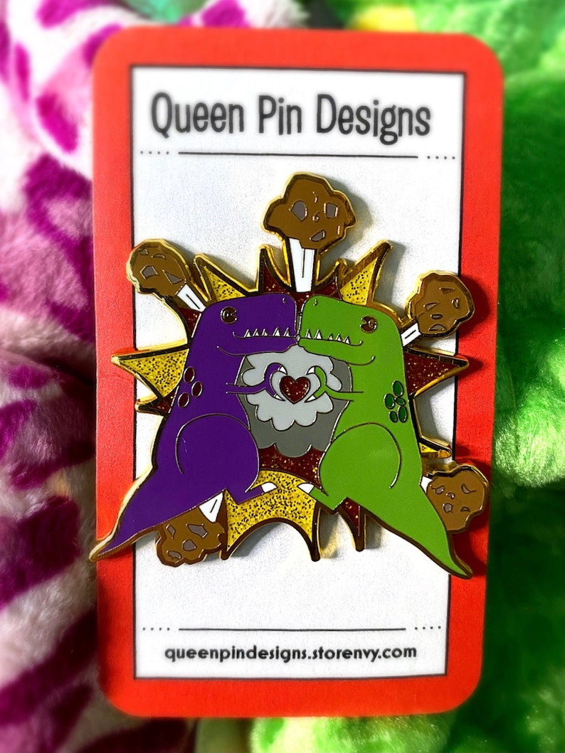 Peut inclure: &Eacute;pingle repr&eacute;sentant deux dinosaures de dessin anim&eacute;, un violet et un vert, s'embrassant dans un motif d'explosion dor&eacute;. L'&eacute;pingle comprend le texte "Queen Pin Designs" et l'adresse queenpindesigns.storenvy.com.