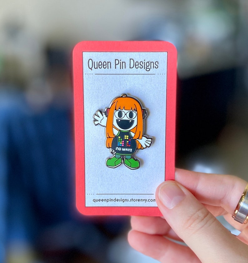Puede incluir: Un colorido pin de esmalte con un personaje de dibujos animados con pelo naranja, auriculares y una camiseta negra con un dise&ntilde;o pixelado. El pin est&aacute; en una tarjeta roja con el texto "Queen Pin Designs". El personaje lleva zapatos verdes y el texto "NO WAY!!" est&aacute; en la camiseta.