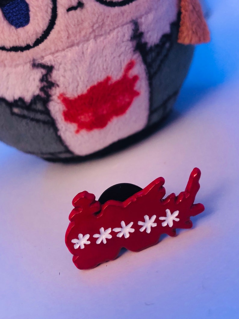 May include: A red enamel pin with white star accents. The pin is in the shape of a stylized word. A plush toy is in the background.