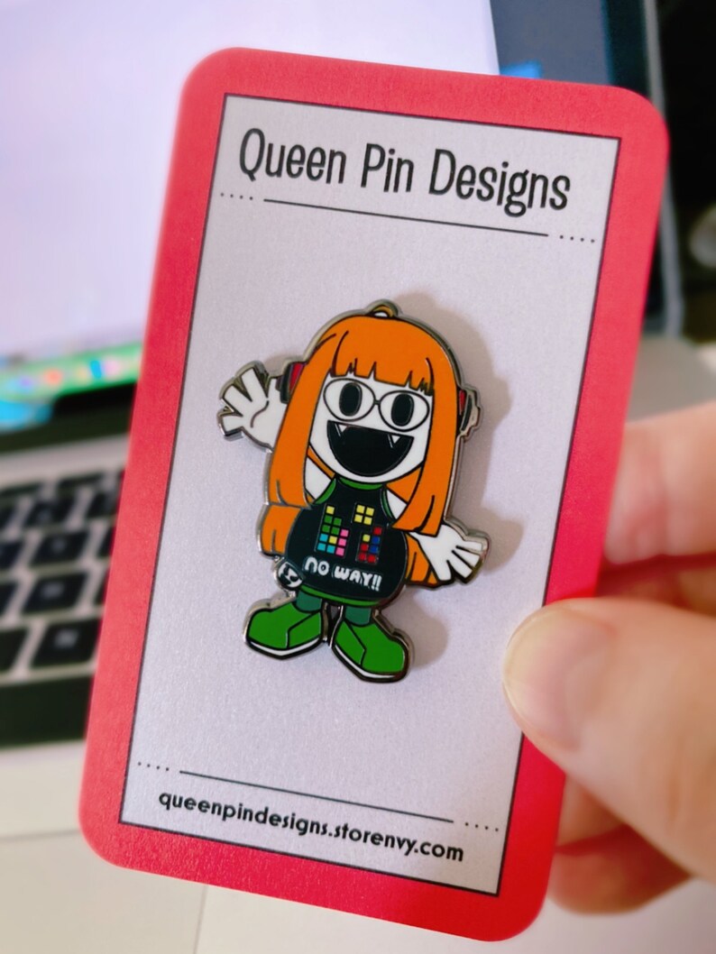 Puede incluir: Un pin de esmalte colorido con un personaje de dibujos animados con pelo naranja, auriculares y un atuendo negro con el texto "NO WAY!!". El pin est&aacute; en una tarjeta con bordes rojos con "Queen Pin Designs" y la direcci&oacute;n del sitio web.
