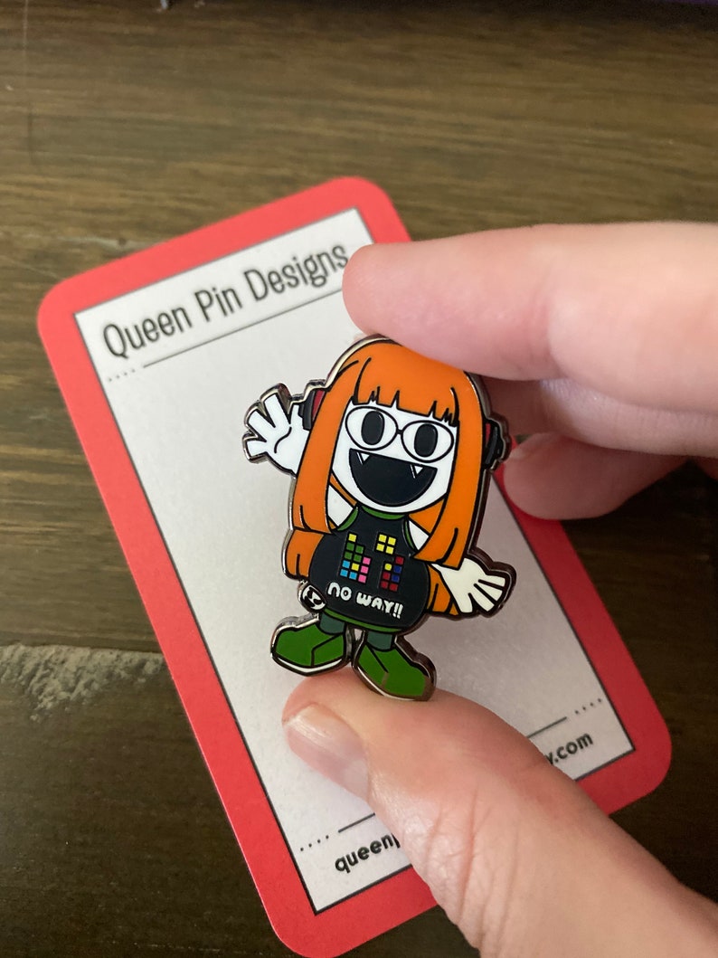 Puede incluir: Un colorido pin de esmalte con un personaje de dibujos animados con pelo naranja, auriculares y una camiseta negra con el texto "NO WAY!!". El pin se muestra sobre una tarjeta con borde rojo con "Queen Pin Designs".
