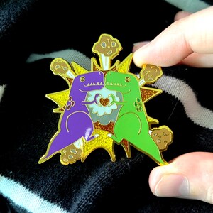 Peut inclure: &Eacute;pingle en &eacute;mail color&eacute;e repr&eacute;sentant deux dinosaures de dessin anim&eacute;, un violet et un vert, se faisant face. Ils sont plac&eacute;s sur un motif d'&eacute;clat jaune et or avec des accents marron. Un c&oelig;ur est au centre.