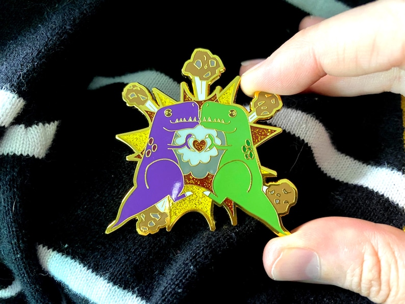 Peut inclure: &Eacute;pingle en &eacute;mail color&eacute;e repr&eacute;sentant deux dinosaures de dessin anim&eacute;, un violet et un vert, se faisant face. Ils sont plac&eacute;s sur un motif d'&eacute;clat jaune et or avec des accents marron. Un c&oelig;ur est au centre.