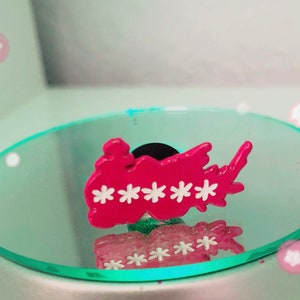 May include: A vibrant pink shoe charm with white star accents. The charm is shaped like a cloud or a stylized shape, with a black backing. The charm is reflected in a mirrored surface.