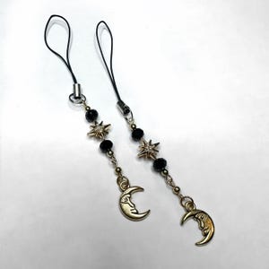 May include: Two celestial-themed phone charms. Each charm features a black cord, black beads, star-shaped charms with rhinestones, and a gold-colored crescent moon pendant. The charms are designed for attaching to a phone or other small device.