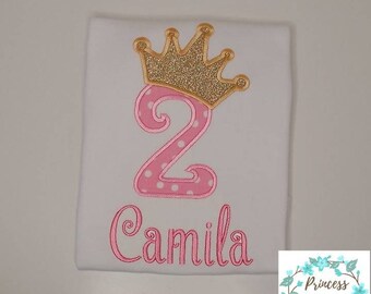 Peut inclure: T-shirt blanc avec un chiffre 2 rose à pois blancs et une couronne dorée à paillettes. Le nom Camila est brodé en rose sous le chiffre. Le t-shirt est pour le 2ème anniversaire d'un enfant.