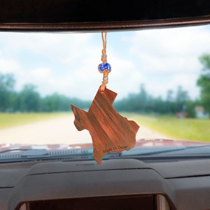 May include: A wooden Texas-shaped car air freshener with a brown wood grain finish and the text "Made in Texas". The air freshener is hanging from a rearview mirror in a car.