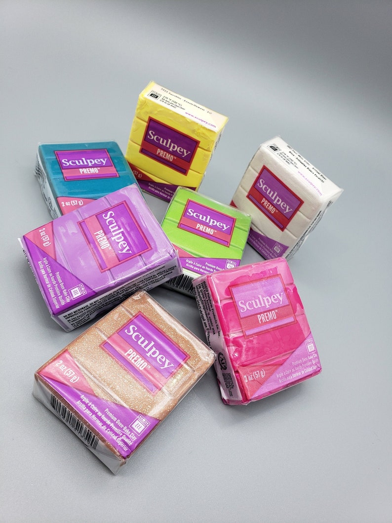 May include: A selection of Sculpey Premo polymer clay blocks in various colours, including yellow, blue, green, pink, purple, white, and gold glitter. Each wrapped block is labelled with the brand name and weight, 57g. Ideal for crafting and modelling.