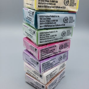 May include: A stack of polymer clay packages in various colours, including yellow, teal, green, pink, purple, white, and brown. Each package has printed text and the AP seal.