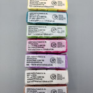 May include: A vertical arrangement of wrapped polymer clay bars in various colours: yellow, teal, green, pink, purple, white, and brown. Each bar has printed labels with manufacturer details and safety information.