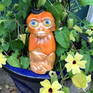 May include: A ceramic monkey figurine with orange fur, blue eyes, and a small black hat. The monkey is set in a blue ceramic pot surrounded by green foliage and yellow flowers.