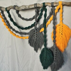May include: A macrame wall hanging with three gray, green, and yellow feather-shaped tassels hanging from a branch. The tassels are made with knotted cords.