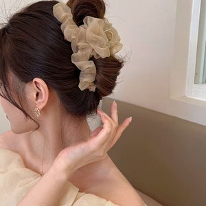 May include: A light brown hair claw with a large, ruffled, sheer fabric bow. The bow is made of a soft, delicate material and is perfect for adding a touch of elegance to any hairstyle.