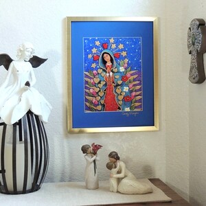 May include: A framed print of a colorful painting depicting the Virgin Mary with a blue background and gold stars. The painting is surrounded by a gold frame and is hanging on a white wall. There are two small white figurines on a table in front of the painting.
