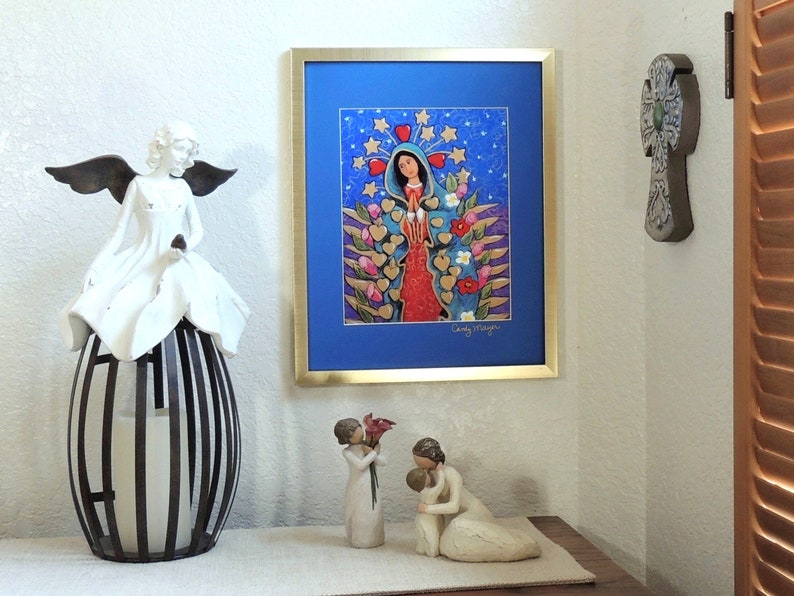 May include: A framed print of a colorful painting depicting the Virgin Mary with a blue background and gold stars. The painting is surrounded by a gold frame and is hanging on a white wall. There are two small white figurines on a table in front of the painting.
