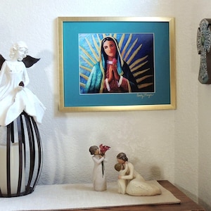 May include: A framed print of a painting depicting the Virgin Mary with a blue robe and a crown of golden rays. The print is hanging on a white wall, with a white angel figurine and two white figurines of a mother and child in the foreground.