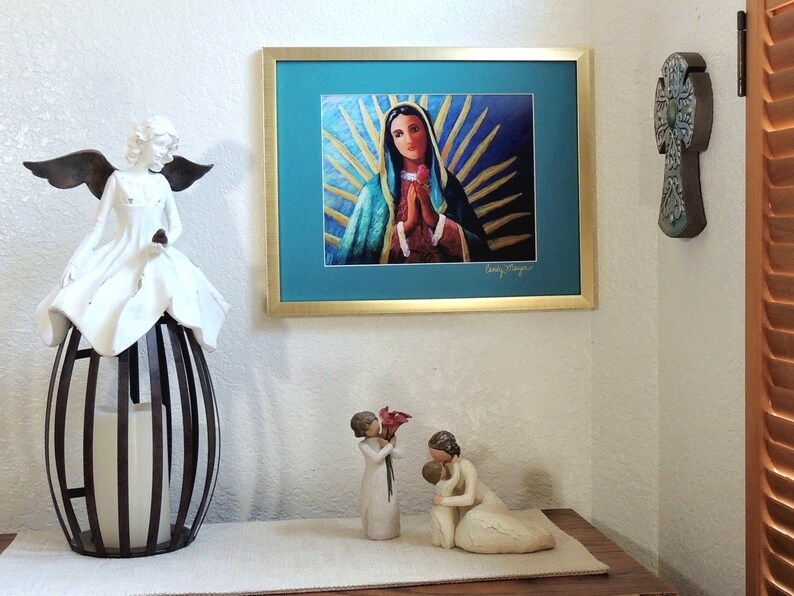 May include: A framed print of a painting depicting the Virgin Mary with a blue robe and a crown of golden rays. The print is hanging on a white wall, with a white angel figurine and two white figurines of a mother and child in the foreground.