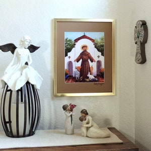 May include: A framed print of a painting depicting Saint Francis of Assisi standing in front of a white building with a brown roof. The saint is wearing a brown robe and holding a bird in his hand. There are other animals in the scene, including a pig, a rabbit, and a cat. The print is on a white wall, and there are two white angel figurines on a table in front of it.