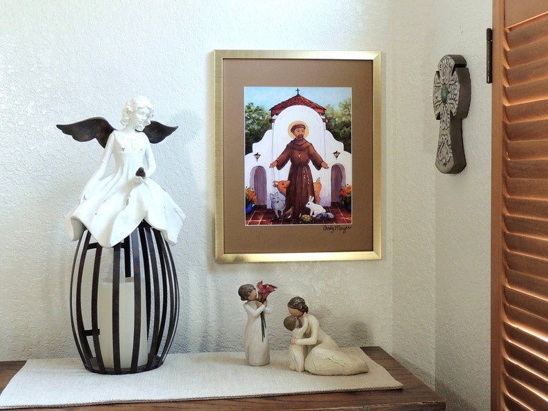 May include: A framed print of a painting depicting Saint Francis of Assisi standing in front of a white building with a brown roof. The saint is wearing a brown robe and holding a bird in his hand. There are other animals in the scene, including a pig, a rabbit, and a cat. The print is on a white wall, and there are two white angel figurines on a table in front of it.