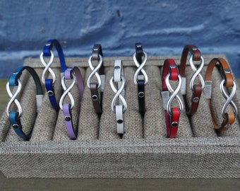Op de afbeelding: Tien leren armbanden met oneindigheidssymbool bedels in verschillende kleuren, waaronder blauw, paars, bruin, wit, zwart, rood en bruin. De armbanden worden getoond op een beige stoffen achtergrond.