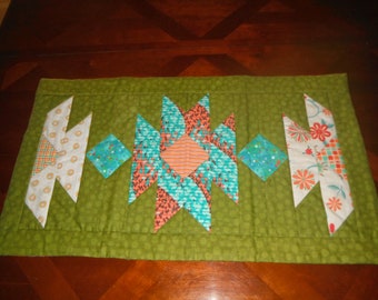 May include: A green table runner with a geometric pattern in shades of orange, teal, and white. The pattern features a central star shape surrounded by diamond shapes.
