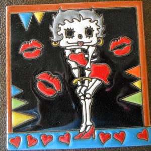 May include: A ceramic tile with a black background and a colorful image of a skeleton wearing a red dress and holding a heart. The skeleton has gray hair and is surrounded by red lips and hearts. The tile has a blue border with yellow, orange, and green triangles.