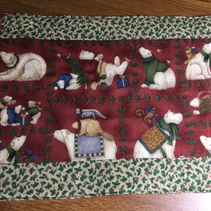 May include: A Christmas-themed table runner featuring polar bears in various poses, some wearing scarves and sweaters. The bears are set against a deep red background with a holly leaf pattern. The edges are trimmed with a green and white holly print.