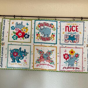 May include: A decorative wall hanging featuring six squares with whimsical illustrations of rhinos and butterflies, surrounded by floral designs. Each square has a positive message, such as "Faithful Friend" and "You be You!" The border is green with a white polka dot pattern.