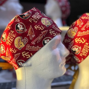 Peut inclure: Bonnets de chirurgien à motifs rouges et dorés avec le logo, le casque et la lance des Seminoles de l'État de Floride. Les bonnets sont présentés sur des têtes de mannequin blanches, mettant en valeur le design et la coupe.