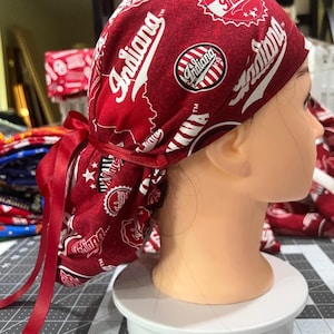 May include: A red and white patterned scrub cap featuring the word "Indiana" in a script font. The cap has a ribbon tie in the back and is displayed on a mannequin head. The pattern includes the outline of the state of Indiana and circular logos.