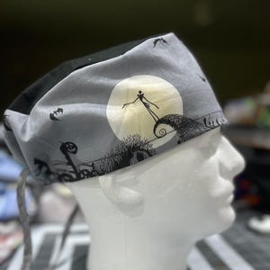 May include: A gray surgical scrub cap featuring a black and white illustration of Jack Skellington from The Nightmare Before Christmas. The design includes a full moon, bats, and a spooky landscape.