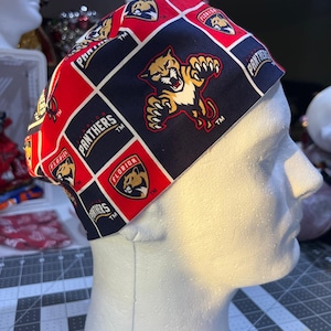 Può includere: Un cappello chirurgico fantasia con il logo dei Florida Panthers. Il cappello ha una combinazione di colori rosso, nero e bianco con il logo e il nome della squadra visualizzati in quadrati. Il cappello è fatto di tessuto ed è progettato per essere indossato in un ambiente medico.