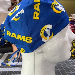 May include: A blue and yellow patterned scrub cap featuring the word "RAMS" and a ram's head logo. The cap is made of fabric and has ties for adjustment. The cap is displayed on a white mannequin head.