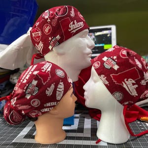 May include: Three burgundy scrub caps with white and red Indiana University logos and text. The caps are on white mannequin heads. The caps have ties in the back.