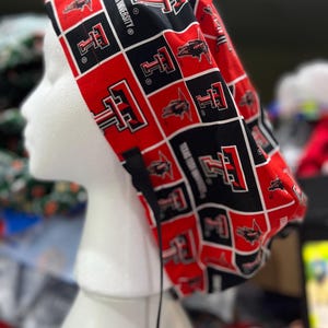 以下が含まれることがあります： 黒と赤の正方形が繰り返されるグリッドパターンのスクラブキャップ。各正方形にはテキサス工科大学のロゴまたは大学名が白い文字で表示されています。キャップは丸みを帯びたギャザー付きの背面と、黒い紐で結ぶ仕様になっています。
