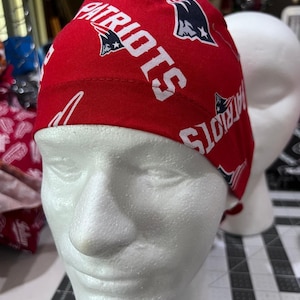 May include: A red and white Patriots patterned cap. The cap features the team's logo and the word "PATRIOTS" in white. The cap is displayed on a white mannequin head.