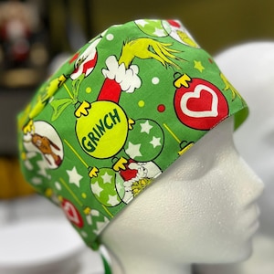 May include: Green scrub cap featuring a festive Grinch-themed pattern. The cap is adorned with Grinch character illustrations, Christmas ornaments, and the word "Grinch" on a yellow ornament. The design includes red, white, and green elements.