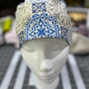 Könnte beinhalten: Eine gemusterte OP-Haube in Blau, Grau und Weiß, dargestellt auf einem Schaufensterkopf. Die Haube hat ein sich wiederholendes Design aus floralen und geometrischen Formen. Der Stoff scheint Baumwolle oder ein ähnliches Material zu sein.