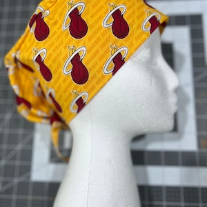 May include: A yellow scrub cap featuring a repeating pattern of the Miami Heat logo in maroon and white. The words "MIAMI HEAT" are printed in white on the yellow background. The cap is tied at the back.