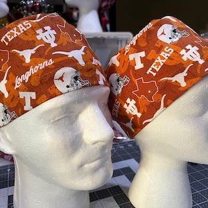 May include: Two orange and white patterned scrub caps. The caps feature the text "TEXAS Longhorns", the letter "T", a Longhorn silhouette, a football helmet, and the outline of the state of Texas.