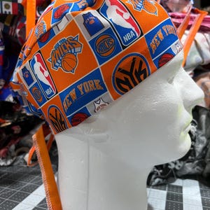 Peut inclure: Une charlotte de bloc opératoire avec un motif orange et bleu vif, arborant les logos des New York Knicks, des graphismes de basket-ball, des emblèmes de la NBA et le texte New York. Elle possède de longs liens en tissu orange à l'arrière.