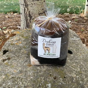 May include: A bag of brown, black, and white alpaca fiber from Dog Leg Ranch in Montana. The bag is tied with twine and has a label that says "Dog Leg Ranch Montana Alpaca's" and an email address.