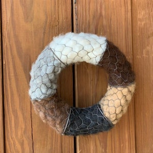 May include: A handmade wreath with a circular design, featuring segments of white, gray, brown, and black wool, encased in a wire mesh. The wreath is hanging on a wooden door.