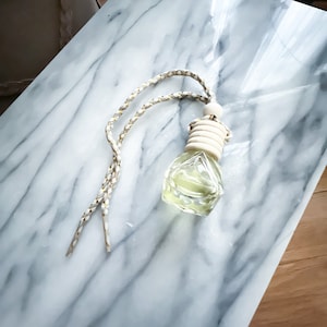 May include: A clear, faceted glass bottle filled with a light green liquid, suspended by a braided white and gold cord. The bottle has a wooden cap and is designed to hang. The background is a marble-patterned surface.