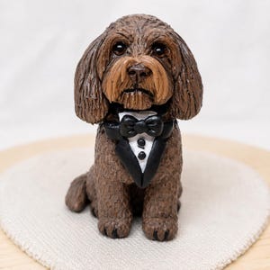 Könnte beinhalten: Eine kleine, braune Hundefigur mit strukturierter Oberfläche, Schlappohren und dunklen Augen. Der Hund trägt ein formelles schwarzes Smoking-Jackett mit weißem Hemdeinsatz und einer schwarzen Fliege. Die Skulptur weist eine detaillierte, lockige Fellstruktur auf.